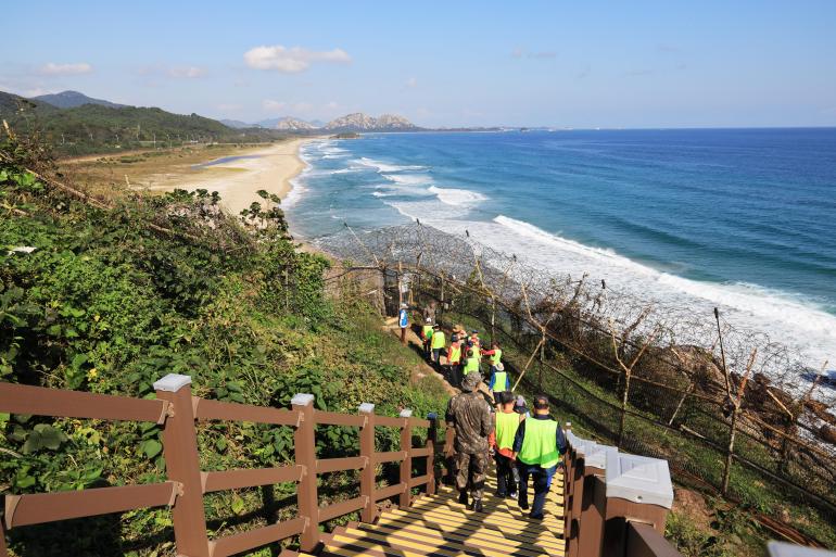 Hikers walking down steps towards the sea. They are wearing reflective shirts. They are walking towards an empty beach with waves crashing onto the shore