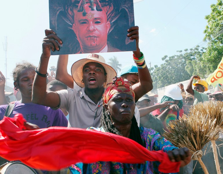 A man in a straw-like hat holds above his head a picture of Laurent Lamothe with horns, red skin and blood dripping from his lips.