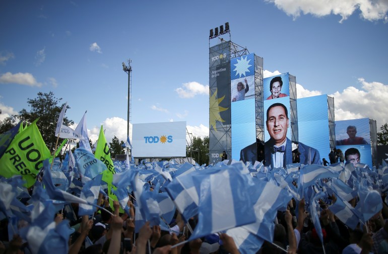 A crowd waving the colors of the Argentinian flag stands below screens showing Juan Peron's face