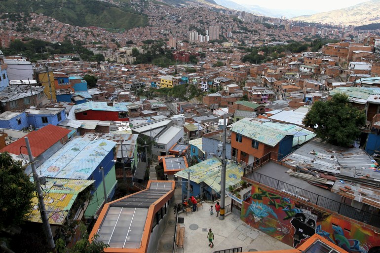 An aerial view of a neighbourhood dominated by tin-roofed buildings, painted orange, red and blue, along a hillside.