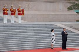 XI Jinping and his wife, Peng Liyuan, on the red carpet at the bottom of the steps outside the Great Hall of the People. There are three soldiers in ceremonial dress half way up the steps behind them.