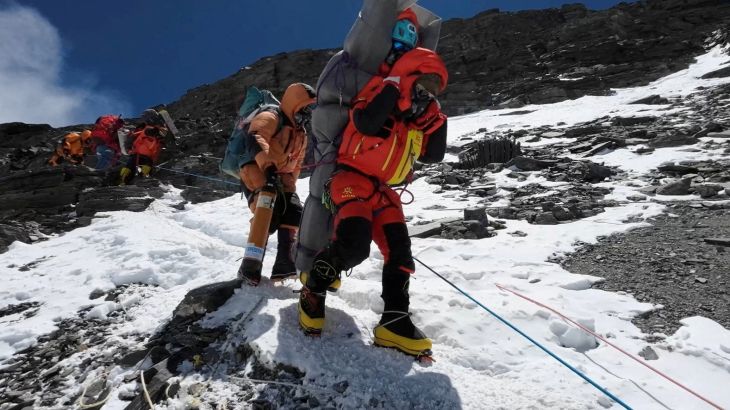 Ngima Tashi Sherpa walks with a Malaysian climber on his back from Mount Everest. It is snowy and rocks are visible. The climber is wrapped in a sleeping mat. Another sherpa is behind.