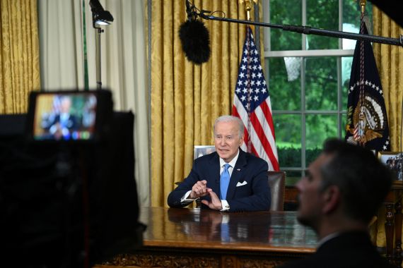Joe Biden, dressed in a dark suit and light blue tie, speaks from behind a heavy wooden desk, surrounded by cameras, monitors and microphones.