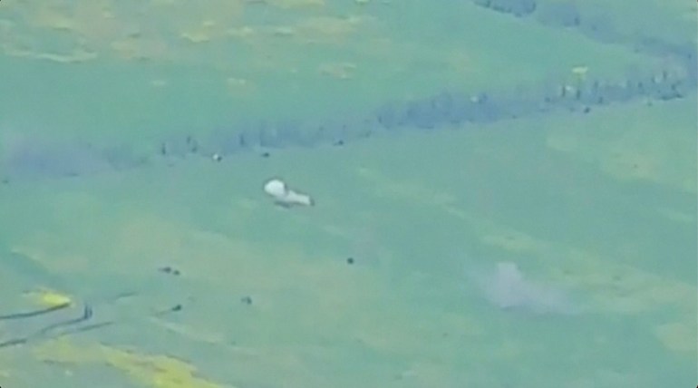 Footage from a drone that Russia says shows armoured vehicles on the move in an undisclosed location. The footage is grainy and shows an expanse of green fields with a number of black dots, and a couple of explosions. There is a row of trees behind.