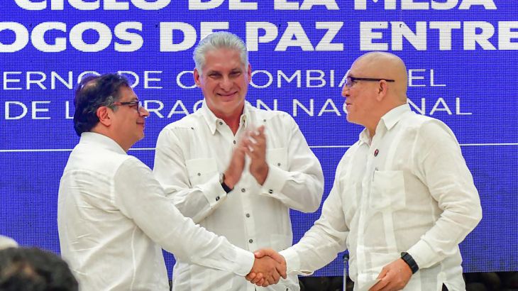 Two men, both dressed in white, shake hands, while another man claps behind them.