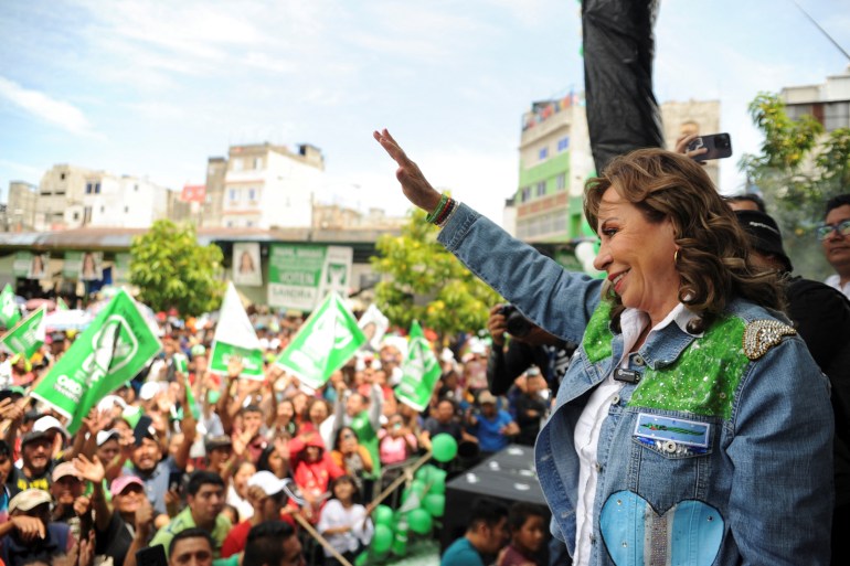 A woman in a blue denim jacket waves to a crowd carrying green-and-white banners.