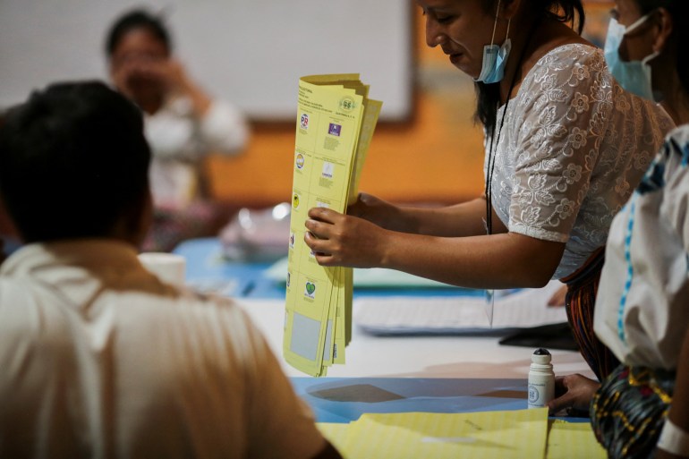 A woman holds a stack of yellow ballots above a ballot box.