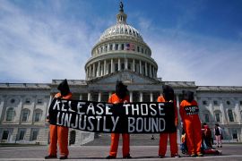 Protesters in orange jumpsuits and black bags over their heads hold sign that reads, "release those unjustly detained"