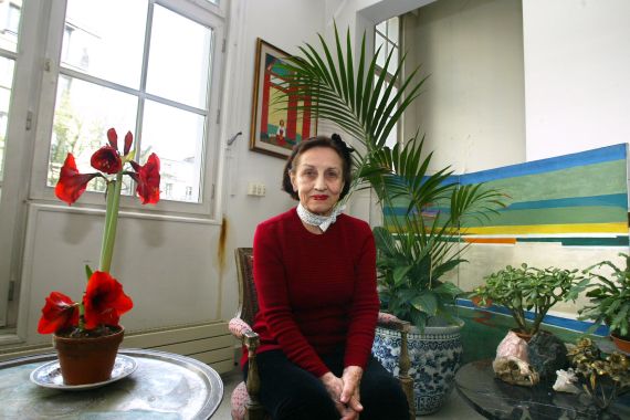 Francoise Gilot picture in her atelier in Paris in 2004. She is wearing a red top and black trousers with a white scarf around her neck, She looks happy and relaxed. There is a vase of red flowers on the table next to her and paintings on the walls around.