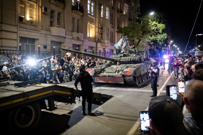 Tanks and soldiers on the street at night time. There are crowds of people on either side of the road taking photos