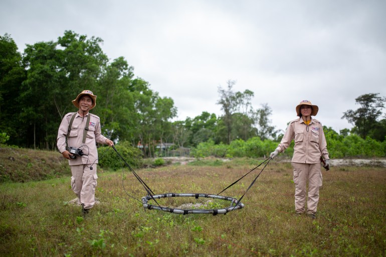 Two men using a loop detector to find UXO. They are wearing the beige uniforms of MAG and walking through a field with the ring between them. There are trees behind. 