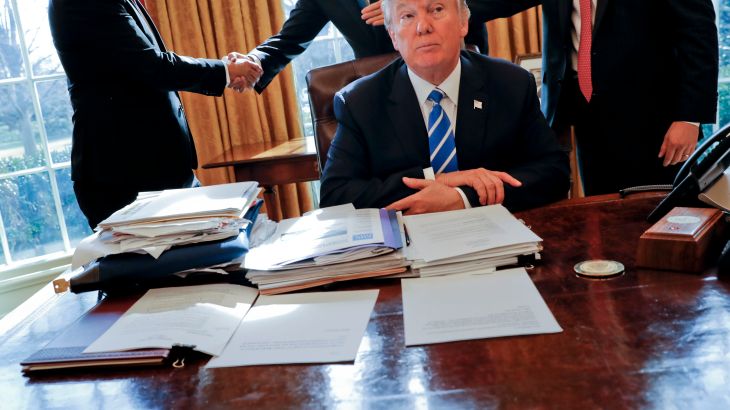 Donald Trump at the Oval Office desk when he was president. There is a pile of documents in front of him and to his right. People are shaking hands behind him.
