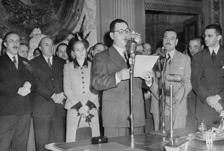 A man in a suit speaks into an old-fashioned microphone, surrounded by dignitaries and with a speech draft in his hand.