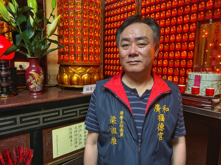 Liang Tsu-wei, honorary chairman of the Guanghe Fude temple. He is standing against a red wall in the temple. He's wearing a blue striped polo shirt, a sleveless jacket in blue with Chinese characters written in yellow. 