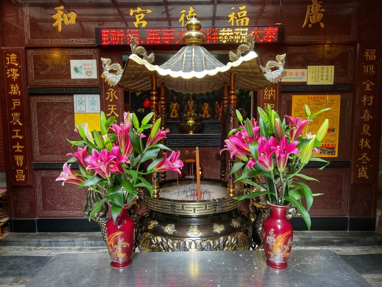 Exterior of the Guanghe Dude temple in Taiwan. There is an urn filled with sand with a joss stick in the middle, with vases of lpink ilies on either side. There is a running LED sign above