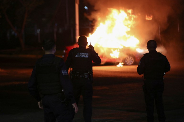 Police officers, seen in silhouette, attend to a car engulfed in flames at night.