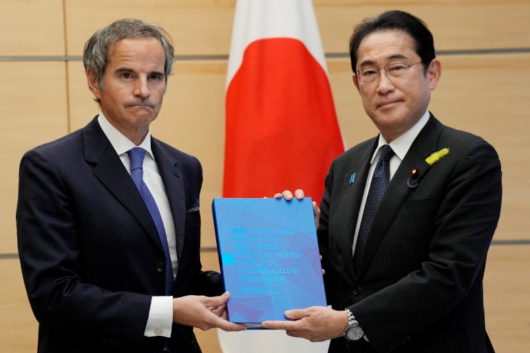 IAEA chief Rafael Grossi handing the report on the Fukushima water release plan to Japanese Prime Minister Fumio Kishida. The report is blue. There is a Japanese flag behind them.