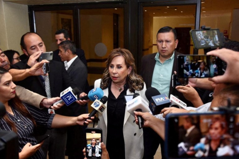 A woman stands surrounded by reporters who hold out microphones to her.