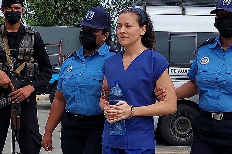 A woman in a blue prison jumpsuit arrives handcuffed with a police escort outside a courthouse. A police van can be seen behind her.