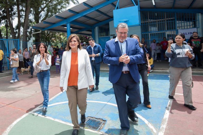 A man in a blue suit walks through an outdoor basketball court with a woman in a white suit jacket and orange shirt, as they prepare to vote.
