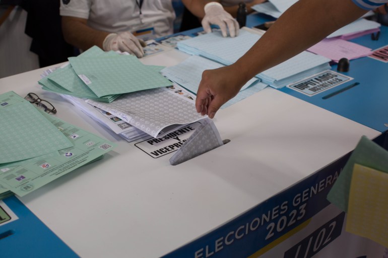 An arm casts a vote into a slot in a large white box.