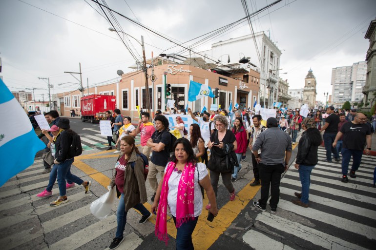 A group of people, some carrying handwritten signs and Guatemalan flags, march across an intersection under a grey, stormy sky.