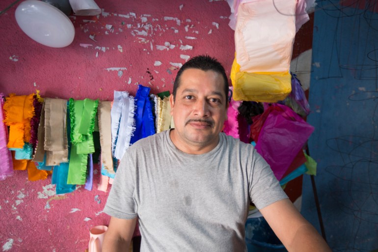 A man in a grey T-shirt stands against a pink wall. A clothesline holds up strips of material for the piñatas.