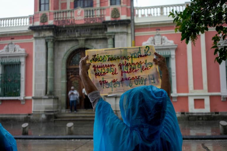 A woman in a blue rain poncho holds up a sign in front of a government building.