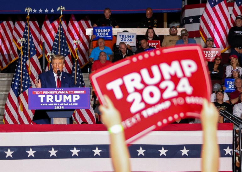 A supporter lifts a "Trump 2024" sign as the candidate speaks from the podium.