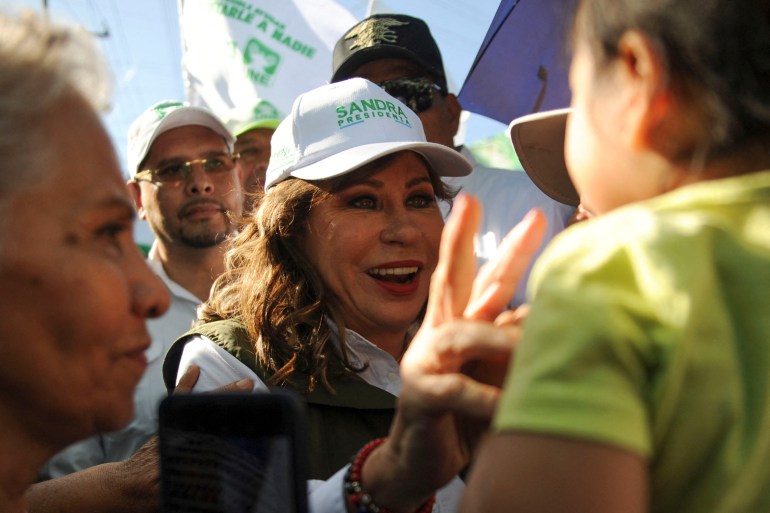 Sandra Torres, wearing a baseball cap emblazoned with "Sandra Presidenta," flashes a peace sign to a young supporter in a crowd.