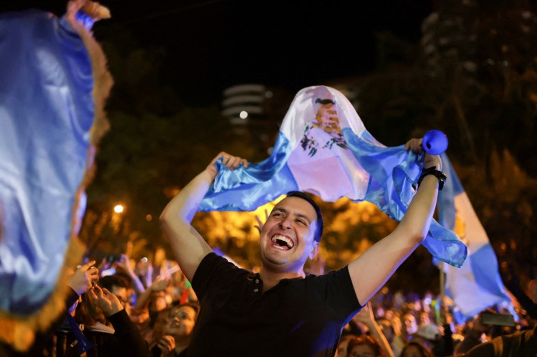 A demonstrator — smiling broadly — waves a Guatemalan flag over his head as he celebrates in a crowded city square.