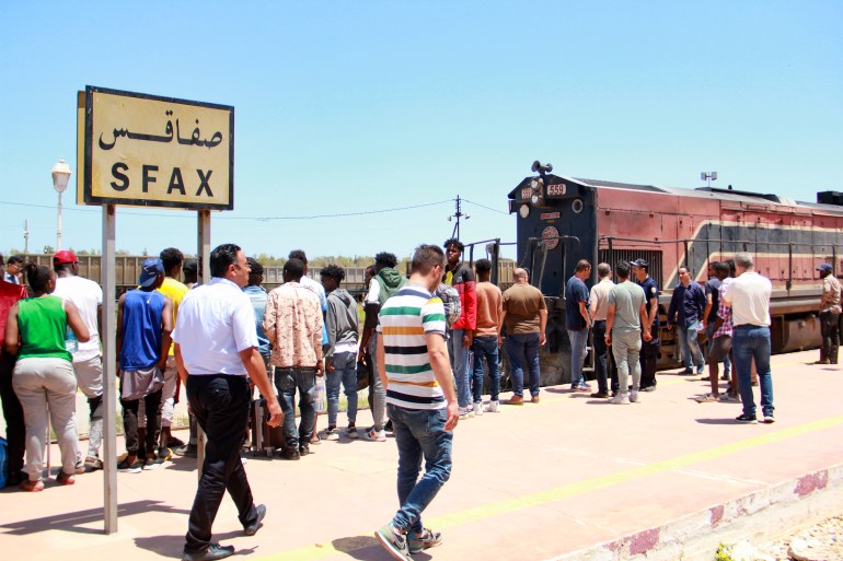 People lining up for a traun outdoors near a sign that reads Sfax in Arabic and French