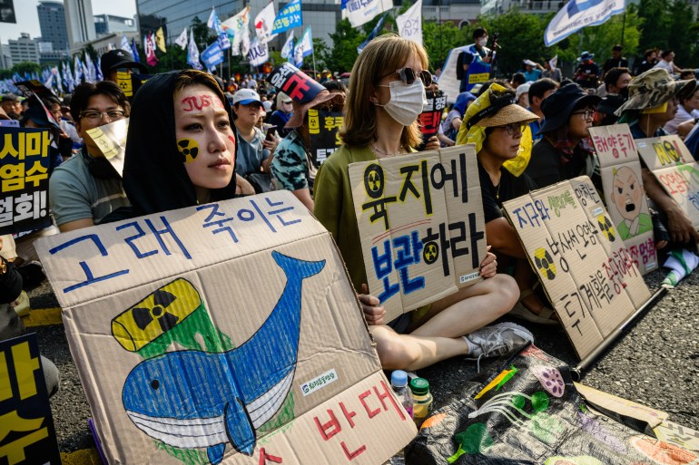 Demonstrators in Seoul show their opposition to the Fukushima discharge plan. They are seated on the ground and one woman has a placard showing a blue and white whale and the symbol for radiation 