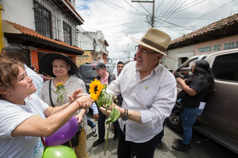 Bernardo Arevalo, wearing a straw hat and carrying a sunflower — a symbol of his campaign —, shakes hands with a supporter on the streets.