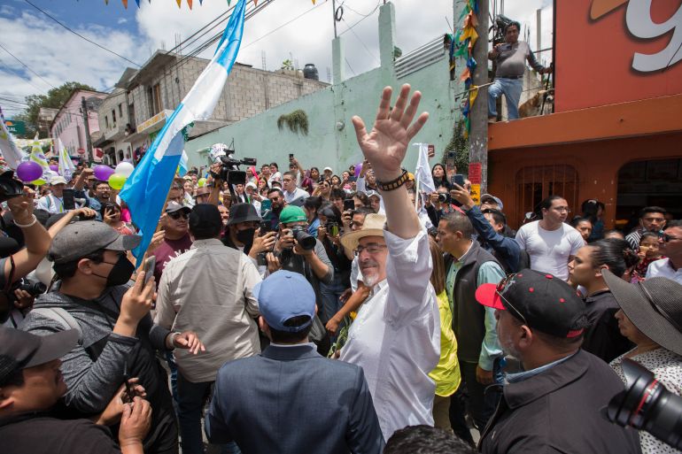 Bernardo Arevalo, wearing a straw hat and a collared shirt, waves from amid a crowd of supporters, one of whom carries a Guatemalan flag.