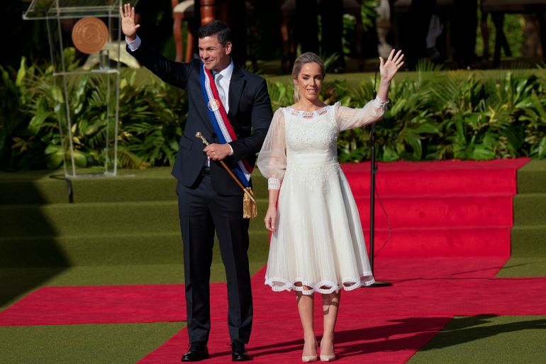 A man in a dark suit and red-white-and-blue sash stands side by side with a woman in a white dress, as they wave from the red carpet.