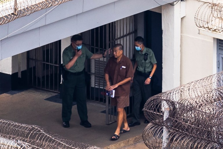 Jimmy Lai being escorted by two prison officers at Hong Kong's Stanley prison. The jail is a maximum security facility and there are coils of barbed wire on the top of the walls. Lai is wearing a brown short sleeved shirt, shorts and slides. 
