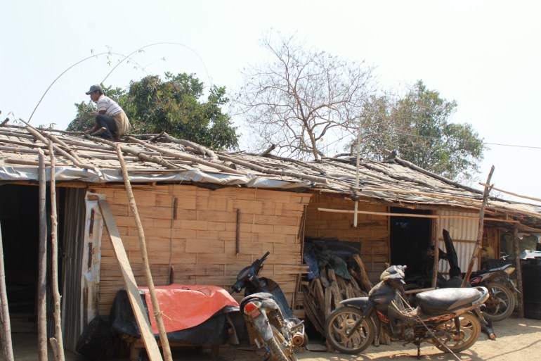 A man is on the roof of shelters made from wood. There are poles leaning against the side of the shelter and some lying on top of it. There are motorbikes parked outside. The man is kneeling on the top of the roof.