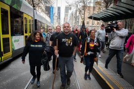 Michael Long (centre) begins his 'long walk' in Melbourne. He is wearing a black t-shirt and jeans and carrying a stick. Other people are walking with him.