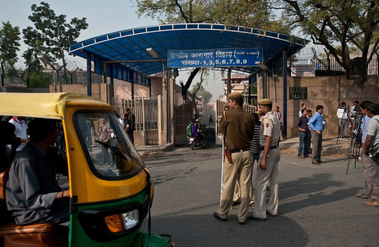 A photo of the outside of the main entrance of Tihar Jail in New Delhi with a tuk tuk on the side and guards and people standing around the gate.