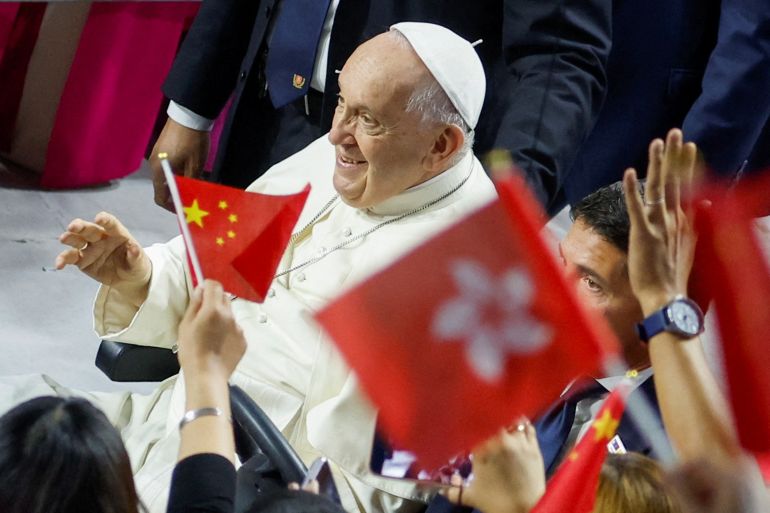 The pope surrounded by Catholic faithful with some waving flags for Hong Kong and China. The pope is smiling.