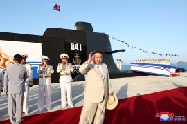 Kim waves at the launch of a new submarine. He is wearing a pale linen suit and carrying a straw hat. The submarine is behind him, It is black with the number 841 written on the side.