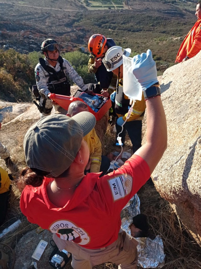 Members of the Mexican immigrant welfare agency Grupo Beta help a person injured during an armed attack by unknown assailants early Friday, at the Cerro de Cuchuma, in Tecate, in Baja California state, Mexico. Wearing a baseball cap and a red shirt, one aid worker lifts a medical packet containing fluids, as other aid workers try to assist a person between two large boulders.
