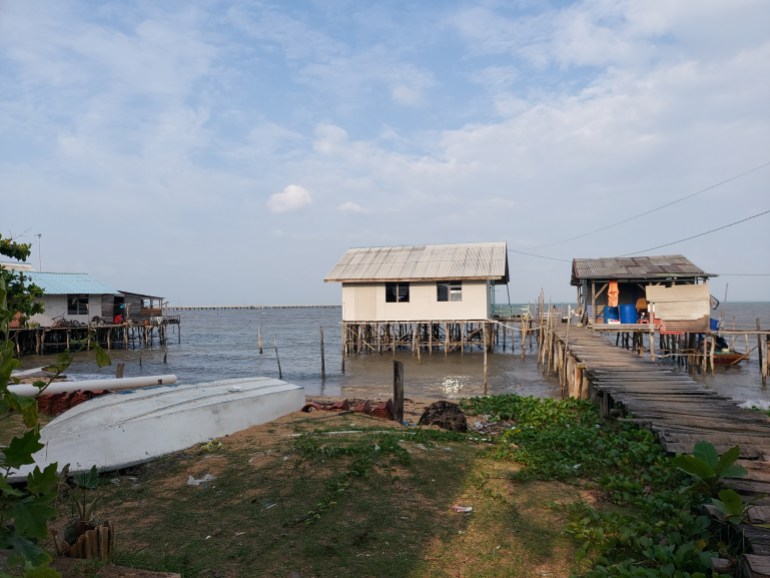Wooden houses on stilts over the sea joined by a wooden walkway. Boats have been hauled up on the shore