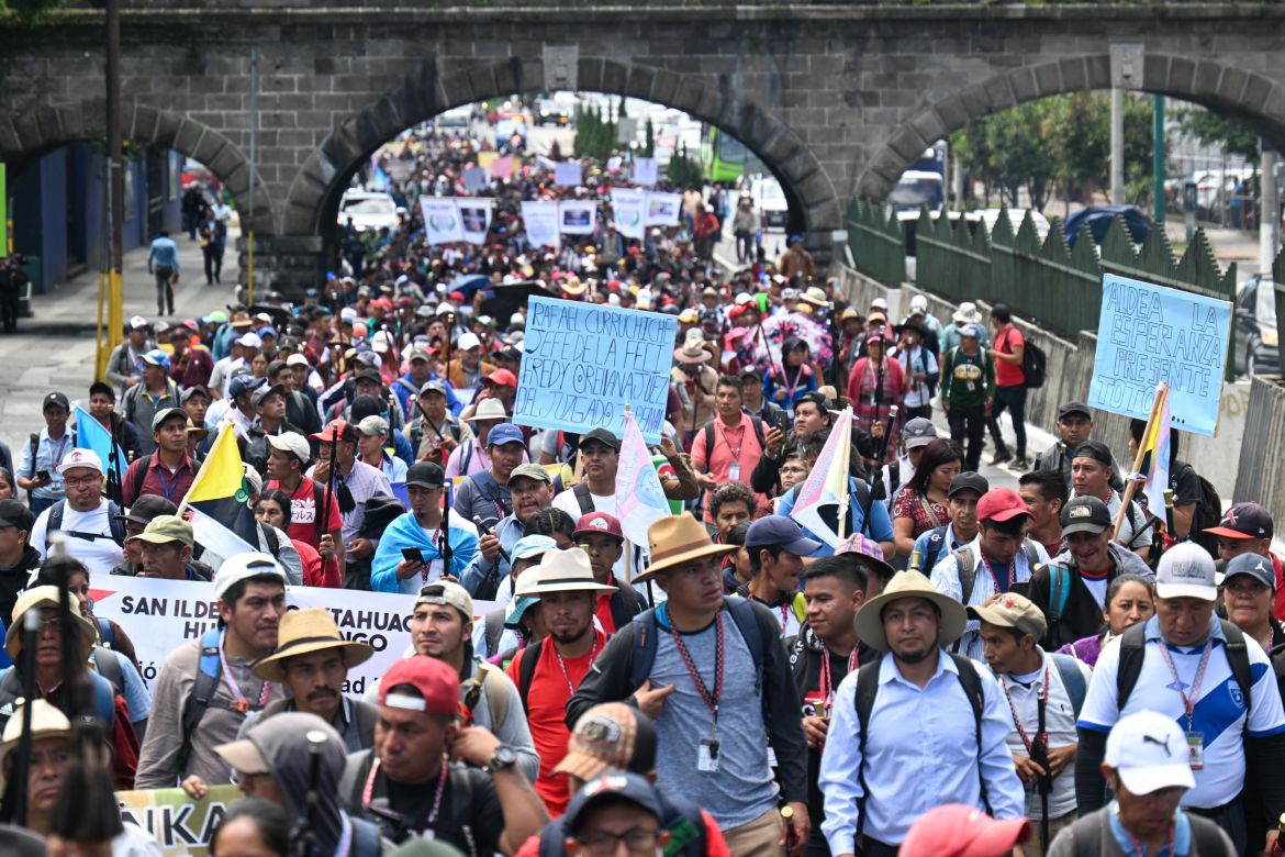 Crowds of people carrying signs file through an archway in Guatemala City.