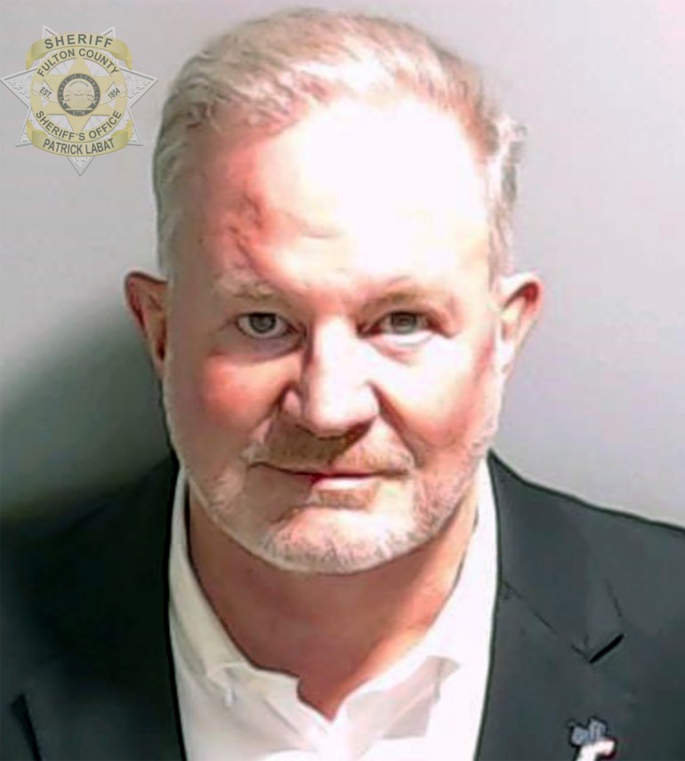 A mugshot of Scott Hall, dressed in a white shirt and dark suit. A Fulton County seal is superimposed at the top of the image.