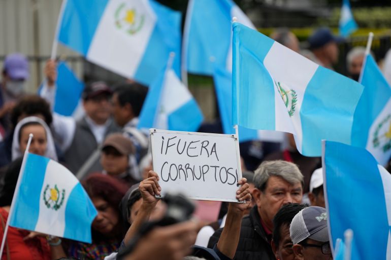 In a sea of protesters waving Guatemalan flags, one protester holds up a handwritten sign that reads: "Fuera Corruptos!"