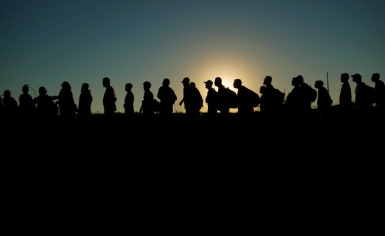 Migrants who crossed the Rio Grande and entered the U.S. from Mexico are lined up for processing by U.S. Customs and Border Protection, Saturday, Sept. 23, 2023, in Eagle Pass, Texas. The line of people are seen in silhouette, shadows against a fading sun.