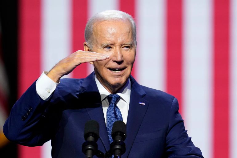 President Joe Biden salutes the crowd as he speaks about democracy and the legacy of the late Arizona Sen. John McCain at the Tempe Center for the Arts, Thursday, Sept. 28, 2023, in Tempe, Ariz. Biden stands in front of a large American flag. His hand is flat in front of his face.