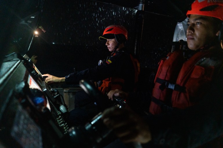 Coast Guard members, dressed in orange helmets and life vests, stand behind the controls of their boat during a nighttime patrol.